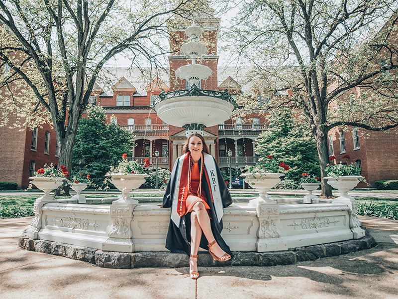 Lauren Grzyboski wearing graduation garb and posing in front of Shippensburg University's Old Main fountain
