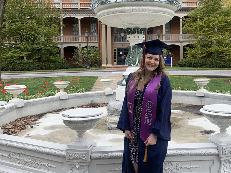 Kirsten Overholtzer wearing cap and gown in front of Shippensburg University's Old Main fountain